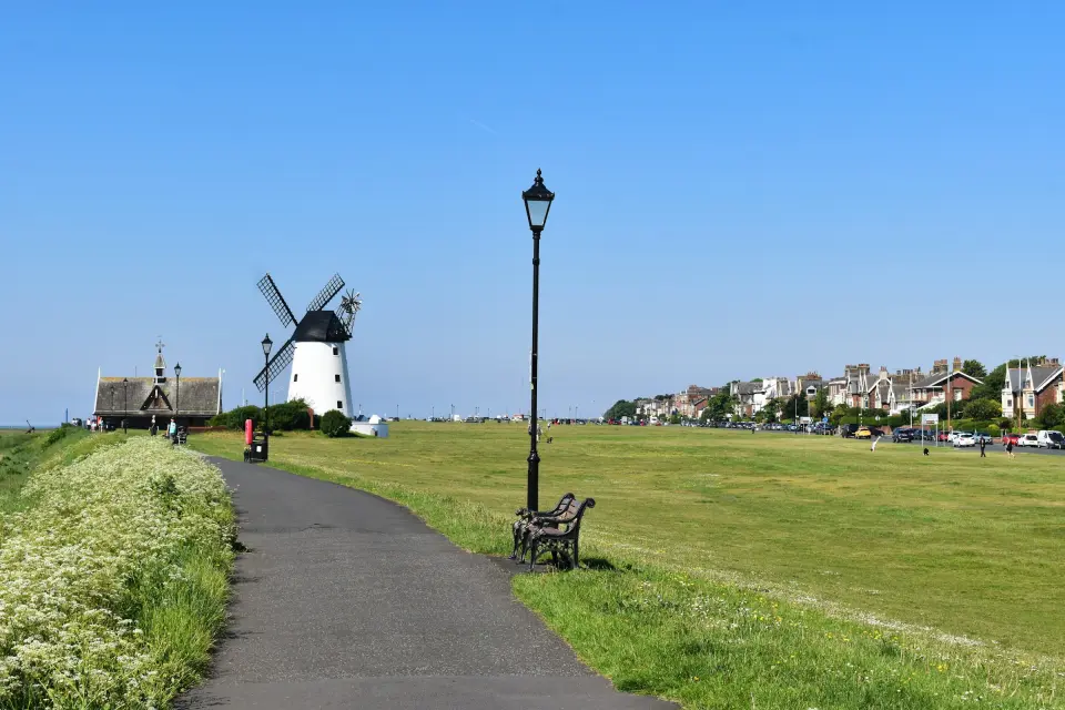 Lytham Windmill and the Old Lifeboat House on Lytham Green, with a footpath, bench and seafront houses under a clear blue sky
